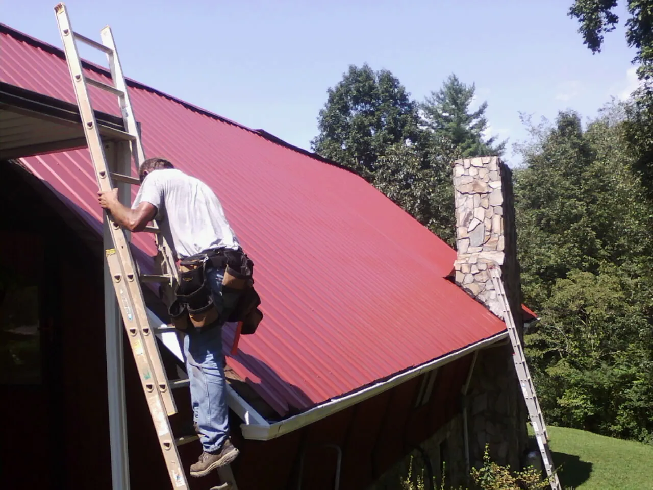 Roofer on a ladder beside a red metal roof and stone chimney — Soffit & Fascia Repair by Roofing Brothers in Spanaway